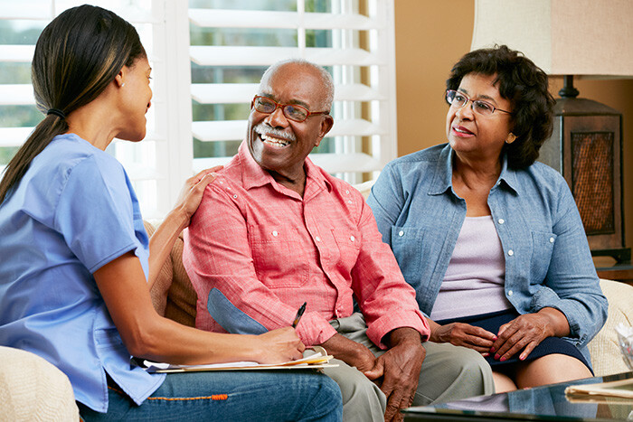 Palliative Care consultation: Nurse in blue scrubs talks with an older couple in a bright living room, smiling and taking notes, representing compassionate healthcare communication and personalized support for seniors.