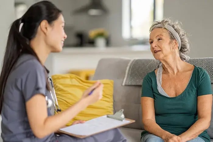Home health conversation: Nurse in gray scrubs speaks with an older woman on a sofa, holding a clipboard, illustrating compassionate in‑home care and patient engagement. Home health conversation: Nurse in gray scrubs speaks with an older woman on a sofa, holding a clipboard, illustrating compassionate in‑home care and patient engagement.