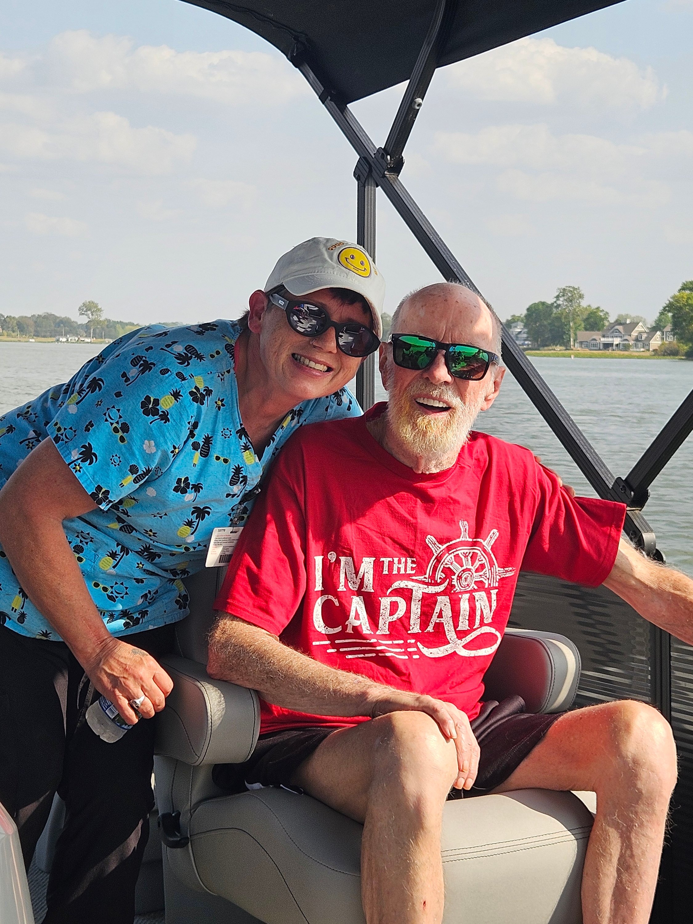 Boating adventure: Smiling older man wearing a red “I’m the Captain” shirt and sunglasses sits at the helm of a boat beside a caregiver in a blue shirt, enjoying a sunny day on the water.