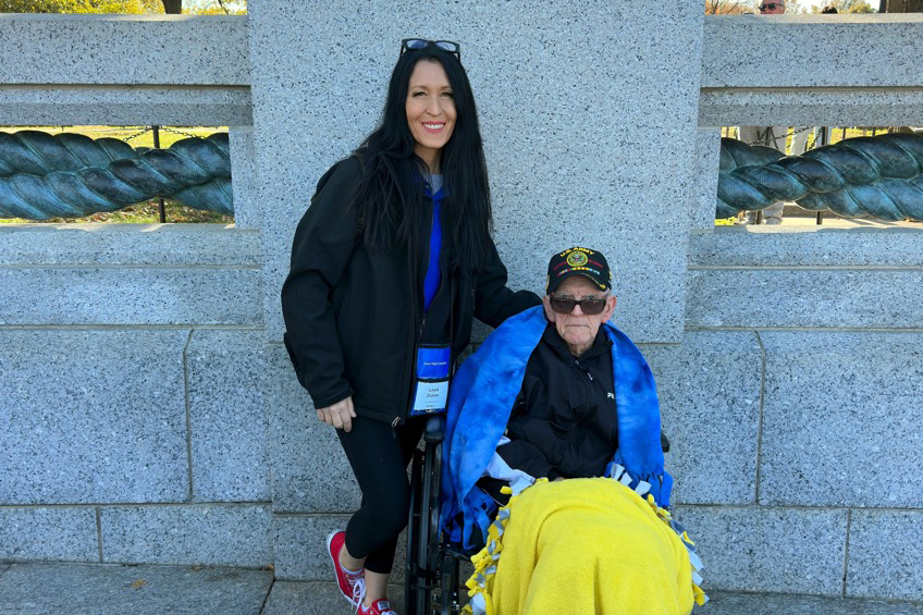 Honor Flight companion and veteran: Woman stands beside an elderly World War II veteran in a wheelchair at a stone memorial. The veteran wears a service cap and is wrapped in blue and yellow blankets, symbolizing gratitude and respect for military service.