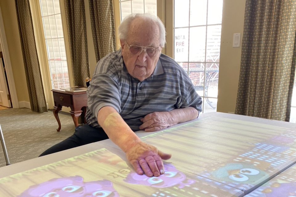 Memory care resident using Lucynt interactive table: Older adult engages with a colorful digital projection featuring animated characters. The technology promotes cognitive stimulation, sensory engagement, and therapeutic recreation in a comfortable senior living environment.
