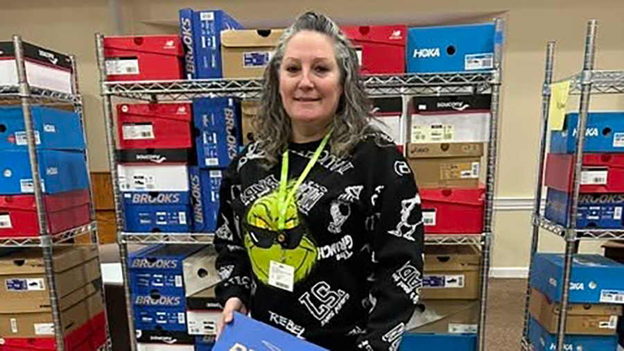 Team member stands in front of shelves stacked with colorful athletic shoe boxes from brands like Brooks, Hoka, and New Balance. She holds a blue box, representing workforce development and support for staff through donated footwear.