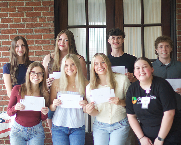 Ohio Living scholarship recipients: Group of seven smiling young adults stand in front of a brick wall holding envelopes of recognition. One wears an Ohio Living shirt, representing community support and educational achievement through senior living scholarship programs.