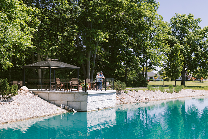 Scenic pondside patio: Two seniors stand on a stone terrace overlooking a clear blue pond surrounded by trees. The shaded seating area with chairs and umbrella offers a peaceful outdoor space for relaxation and community connection at a senior living campus.