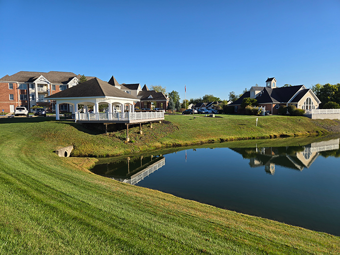 Ohio Living Cape May campus featuring a scenic pond surrounded by manicured green lawns and walking paths. A white gazebo with railing overlooks the water, reflecting alongside brick and siding buildings under a clear blue sky. The peaceful outdoor setting highlights the community’s serene environment and well‑maintained landscape for senior living.