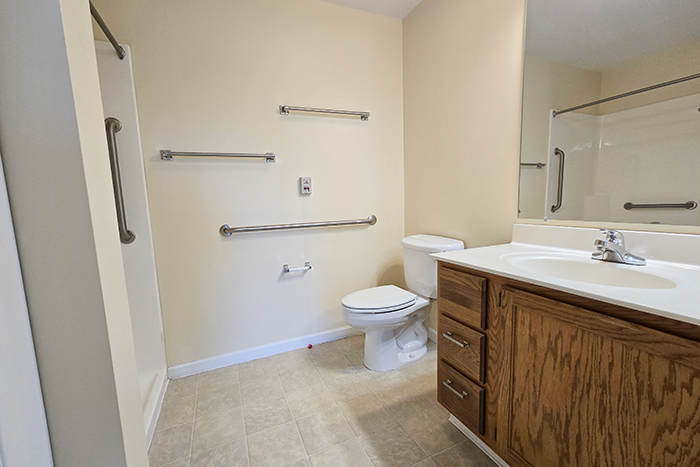 Ohio Living Cape May Cambridge Apartments bathroom featuring an accessible design with beige walls, light tile flooring, and a walk‑in shower with stainless steel grab bars. The space includes a toilet with multiple safety rails, a wooden vanity with white countertop and sink, and a large mirror, providing a safe and comfortable environment for senior independent living.