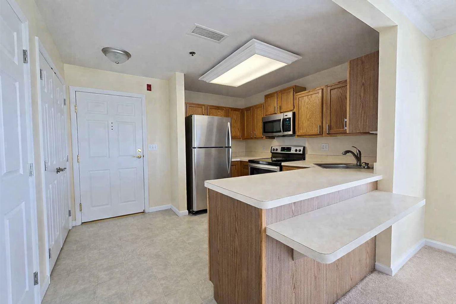 Ohio Living Cape May Cambridge Apartments kitchen featuring light beige walls, tile flooring, and oak cabinetry. The space includes stainless steel appliances—refrigerator, oven, and microwave—plus a double sink and extended countertop with breakfast bar. Bright overhead lighting and neutral finishes create a modern, accessible design for senior independent living.