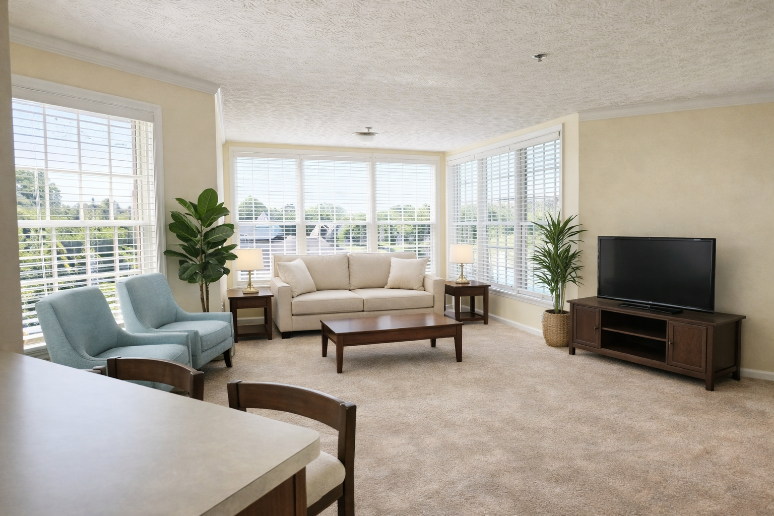 Ohio Living Cape May Cambridge Apartments living room featuring large corner windows with white blinds, beige carpet, and neutral walls. The space includes a cream sofa, two light blue armchairs, wooden coffee and side tables with lamps, a flat‑screen TV on a wooden stand, and potted plants adding greenery. Natural light fills the room, creating a bright, comfortable environment for senior independent living.