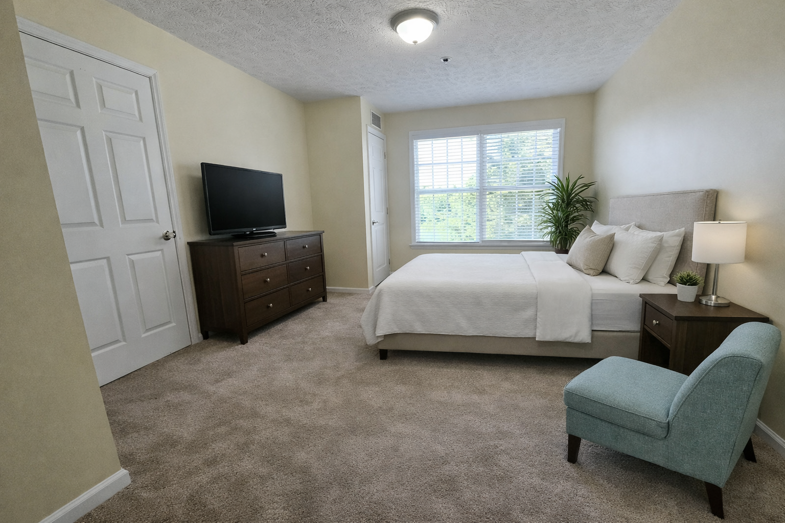 Ohio Living Cape May Cambridge Apartments bedroom featuring a queen bed with white linens, beige walls, and soft carpet flooring. The room includes a wooden nightstand with lamp and plant, a matching dresser with flat‑screen TV, and a light blue upholstered chair. Large windows with white blinds fill the space with natural light, creating a bright, comfortable environment for senior independent living.