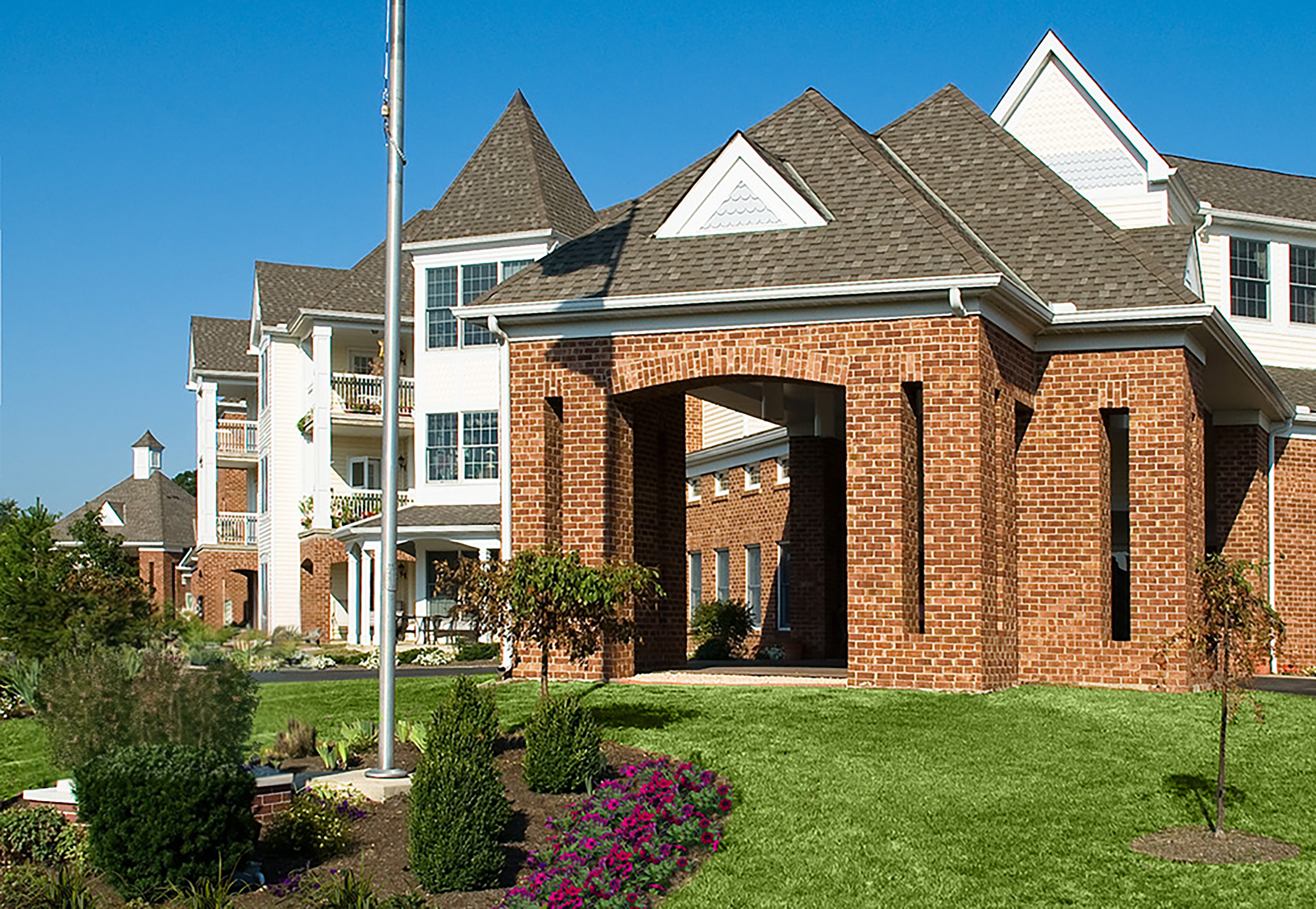 Ohio Living Cape May Cambridge Apartments featuring a multi‑story brick and white‑sided building with gabled roofs and large windows. The structure includes a covered brick entrance, balconies, and landscaped grounds with green lawns and shrubs under a clear blue sky.