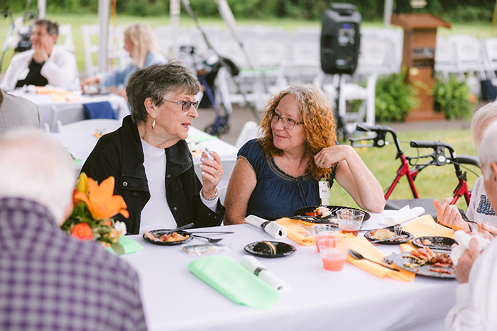 Two women seated at a table outdoors during a community luncheon at Ohio Living Cape May engage in friendly conversation. The table is decorated with bright orange and green napkins and a floral centerpiece featuring orange lilies. Plates of food and drinks are visible, with other attendees and event seating in the background under a tent. The scene conveys warmth, connection, and active senior living in a welcoming environment.
