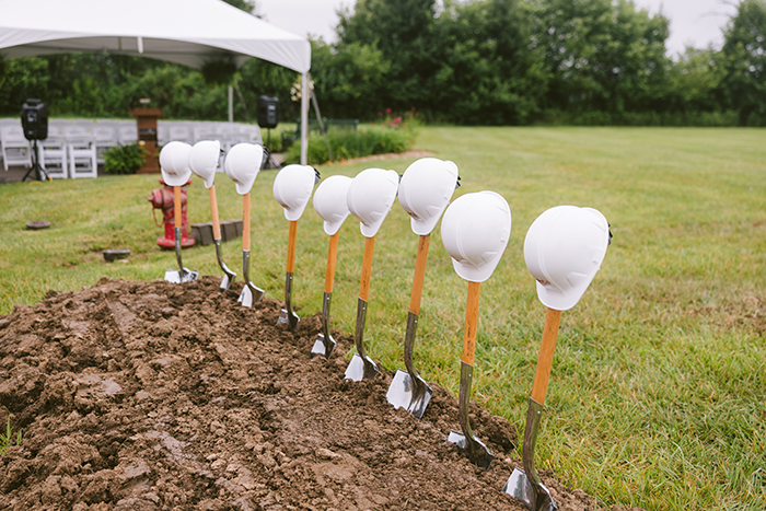 A row of nine shovels with wooden handles and shiny metal blades stand upright in freshly turned soil, each topped with a white construction hard hat. The setup is part of a ceremonial groundbreaking event at Ohio Living Cape May, symbolizing the start of new development for the senior living community. In the background, a white canopy tent with chairs and speakers sits on a grassy field surrounded by trees, representing progress and community growth.
