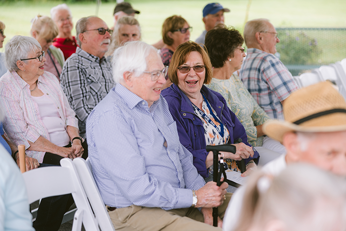 Residents and guests at Ohio Living Cape May sit together under a canopy during a community event. The group of older adults, dressed casually, smile and chat while seated in white folding chairs arranged in rows. The outdoor setting features green grass and trees in the background, creating a warm, welcoming atmosphere that reflects social engagement and active senior living.