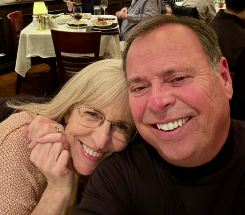 A close-up photo of two smiling adults seated together at a restaurant. One person rests their head affectionately on the other’s shoulder while they pose for the camera. Tables, diners, and soft lighting create a warm and welcoming dining atmosphere.