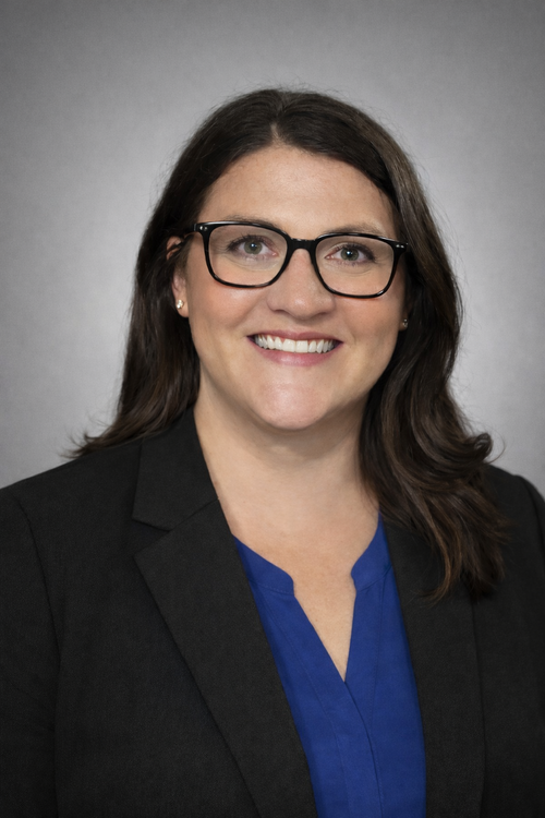 A professional headshot of a woman with dark hair and glasses, wearing a black blazer over a blue blouse. She is smiling against a neutral gray background, creating a polished and approachable business portrait.