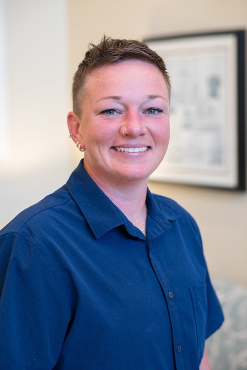 A professional portrait of a smiling person with short hair, wearing a blue collared shirt. They are photographed indoors with a softly blurred background, creating a warm, approachable, and welcoming appearance. The image conveys confidence, friendliness, and professionalism.