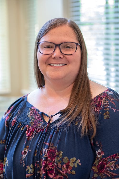 A professional portrait of a smiling woman with glasses and long brown hair, wearing a blue floral blouse. She is photographed indoors with soft natural light and window blinds in the background, creating a warm and welcoming appearance.
