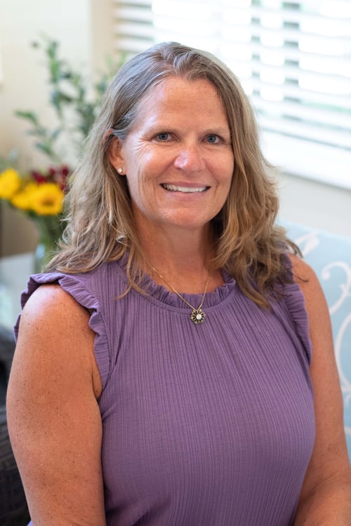A smiling woman in a lavender top is seated indoors with soft natural light and flowers in the background. Her warm expression and relaxed posture create a welcoming, friendly presence that reflects compassion, confidence, and approachability.