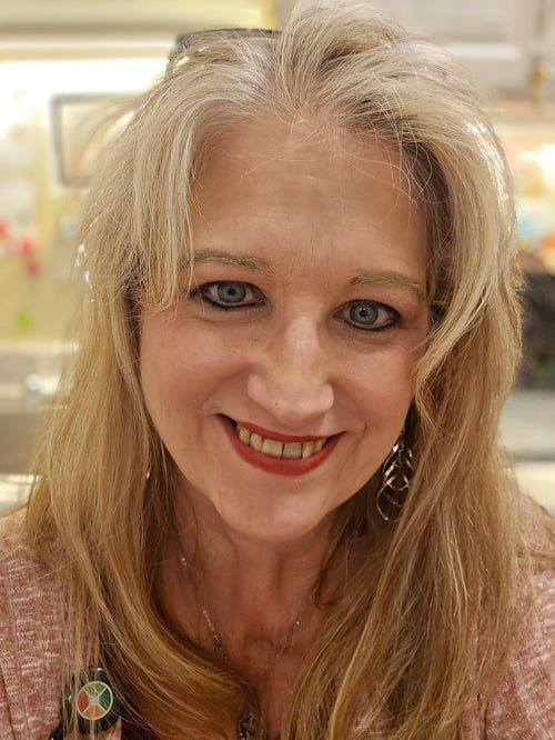 A smiling woman with long blonde hair is pictured indoors in a warmly lit setting. Her friendly expression, bright eyes, and welcoming smile create a sense of kindness, compassion, and approachability.