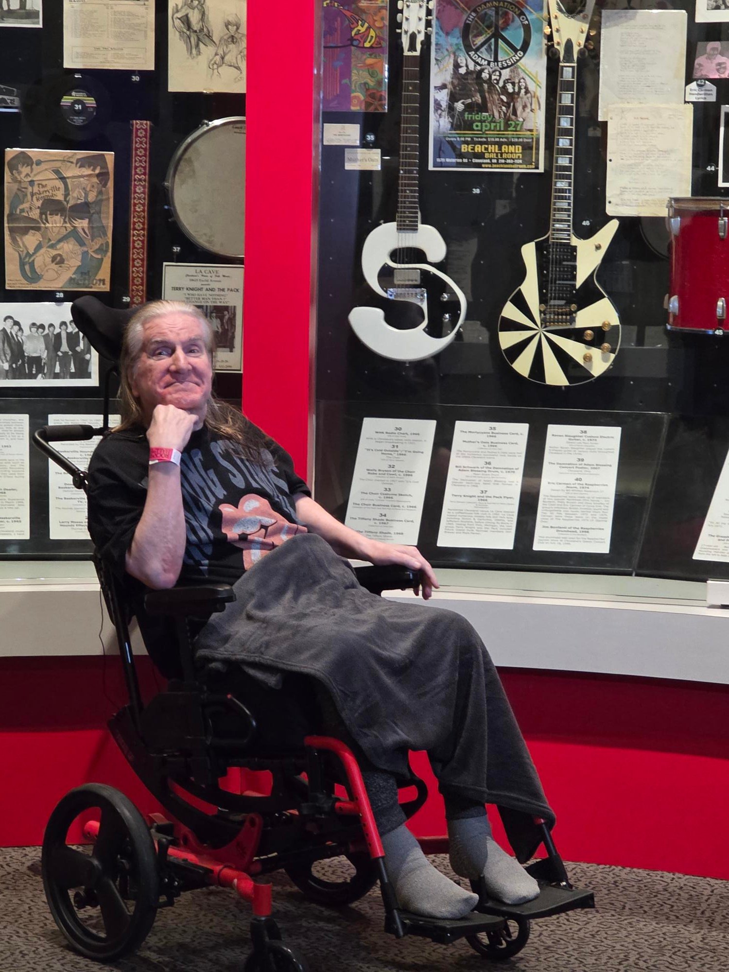 Ohio Living hospice patient at the Rock &amp; Roll Hall of Fame exhibit: Older man in a wheelchair smiles beside a display of guitars, posters, and music memorabilia, celebrating rock history and accessible cultural experiences for seniors.