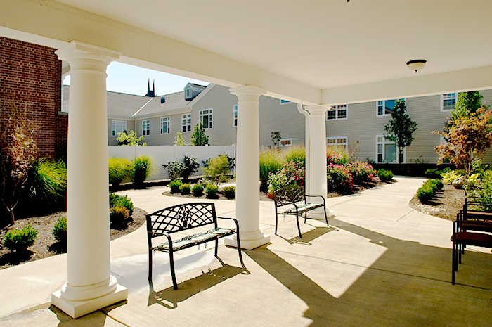 Covered outdoor courtyard at Ohio Living Mount Pleasant featuring white columns, decorative benches, and landscaped garden beds with shrubs, flowers, and ornamental grasses. The bright, sunny space is surrounded by residential-style buildings, offering a peaceful and secure environment designed for memory care residents to relax and enjoy nature.