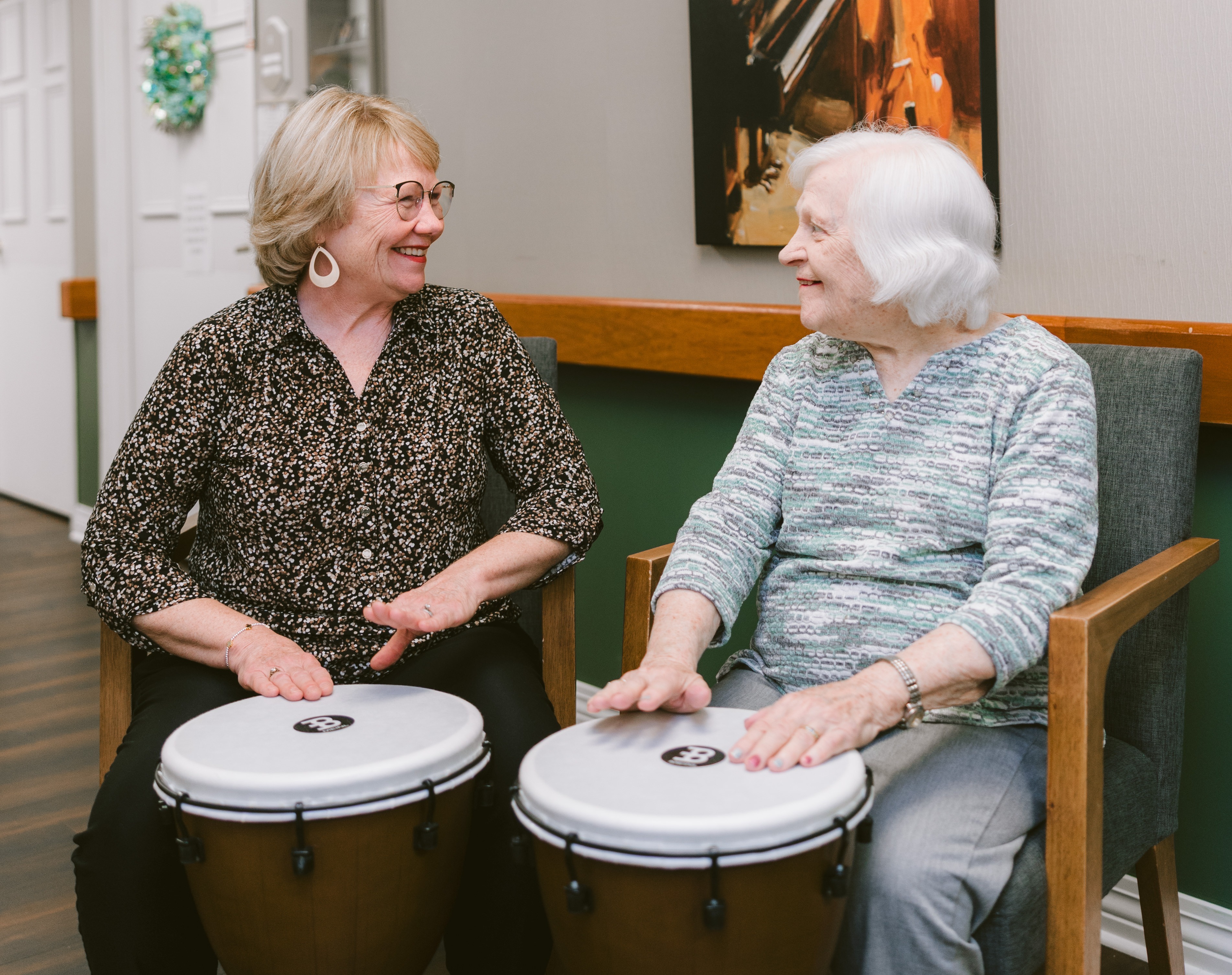Two women share a joyful moment during a drumming activity, smiling and engaging with one another. The image highlights the power of music, connection, and meaningful social interaction in creating a vibrant and enriching community experience.