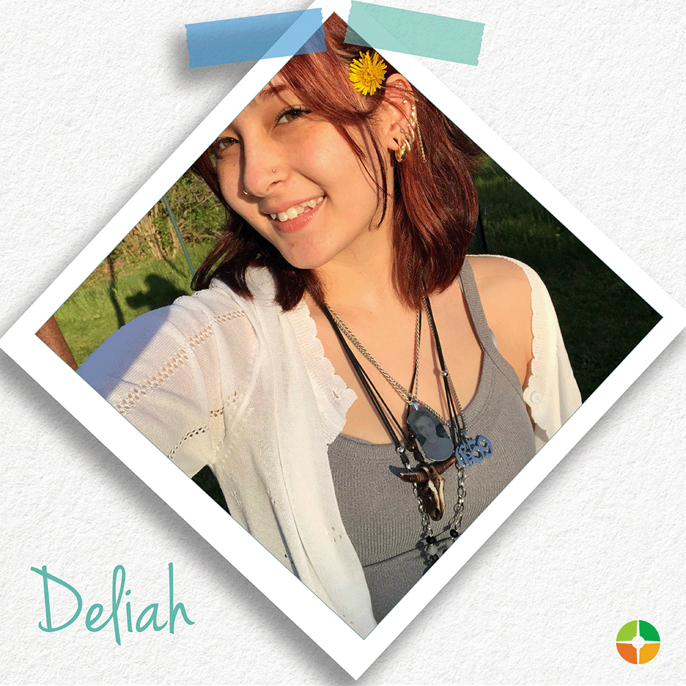 Polaroid-style selfie of a smiling young woman with red hair outdoors in sunlight. She wears layered necklaces, earrings, and a light sweater over a gray top, with a yellow flower tucked in her hair. The name “Deliah” appears in teal script at the bottom, with a small logo in the corner.