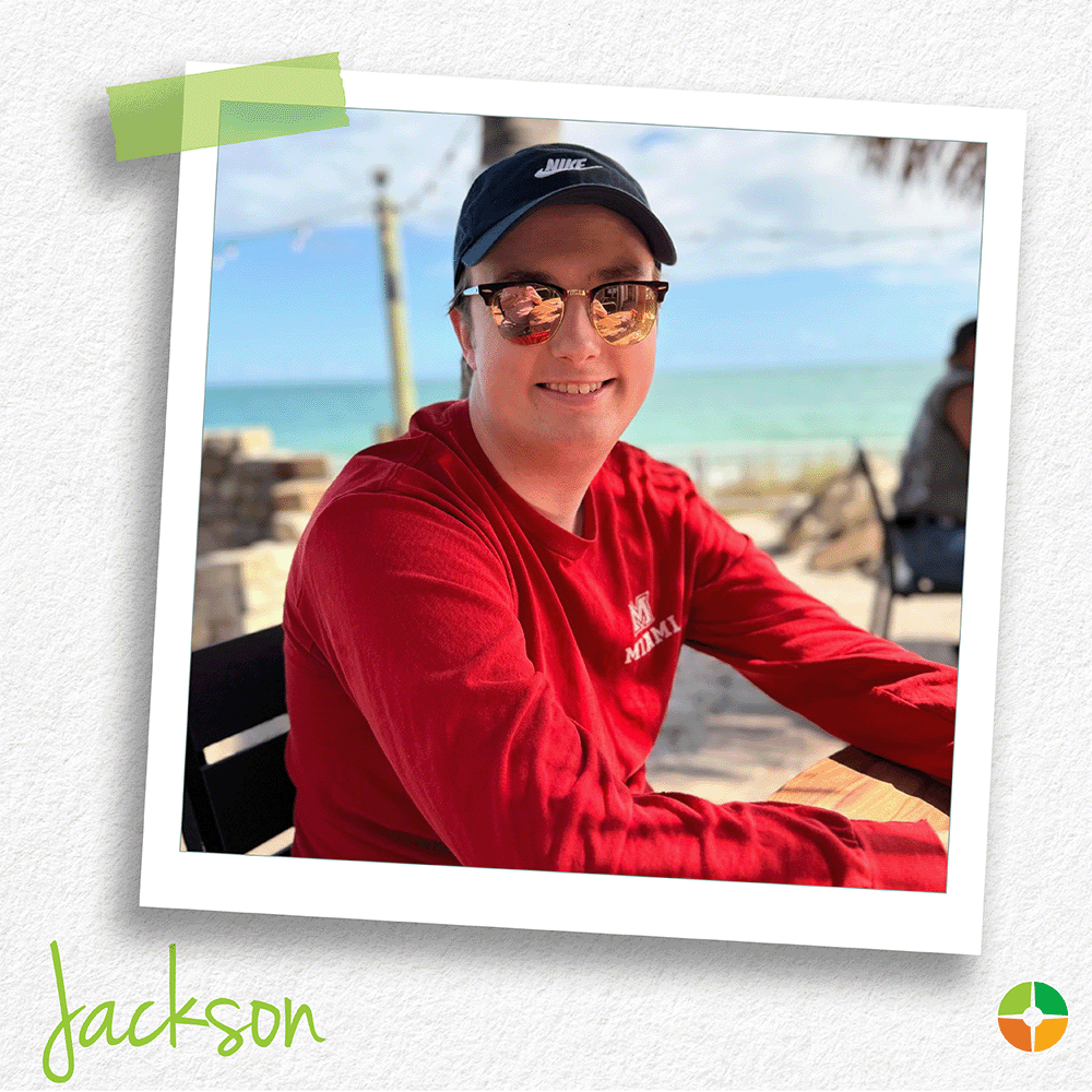 A senior portrait-style photo of Jackson seated at an outdoor beachside table. Wearing a red shirt, sunglasses, and a cap, he smiles toward the camera with the ocean and shoreline visible in the background. A senior portrait-style photo of Jackson seated at an outdoor beachside table. Wearing a red shirt, sunglasses, and a cap, he smiles toward the camera with the ocean and shoreline visible in the background.