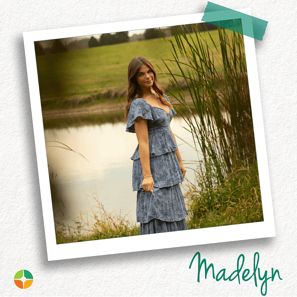 Polaroid-style photo of a young woman standing beside a pond in a grassy outdoor setting. She wears a layered blue dress and looks toward the camera. Tall reeds and water appear in the background. The name “Madelyn” appears in green script at the bottom, with a small logo in the corner.