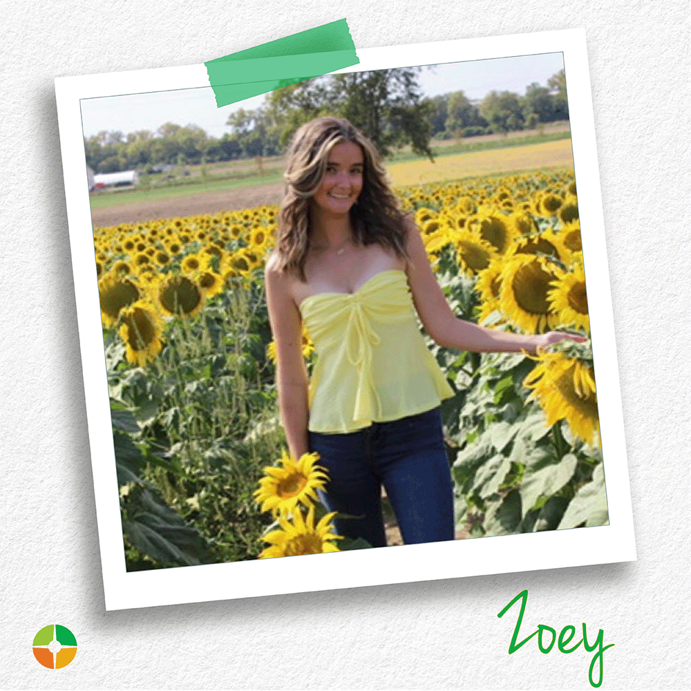 Polaroid-style photo of a smiling young woman standing in a sunflower field on a sunny day. She wears a light yellow top and jeans and holds a sunflower. The name “Zoey” appears in green script at the bottom, with a small logo in the corner.