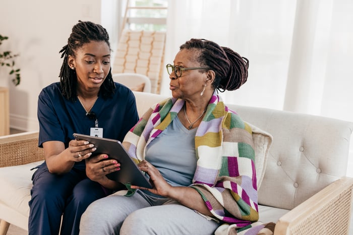 Home health consultation: Healthcare professional in navy scrubs assists an older woman using a tablet on a sofa, representing personalized home health support and digital care coordination. Home health consultation: Healthcare professional in navy scrubs assists an older woman using a tablet on a sofa, representing personalized home health support and digital care coordination.