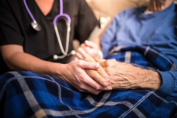 Hospice care compassion: Nurse in black scrubs gently holds the hand of an elderly patient resting under a blue plaid blanket, symbolizing comfort, dignity, and compassionate end‑of‑life care at home.