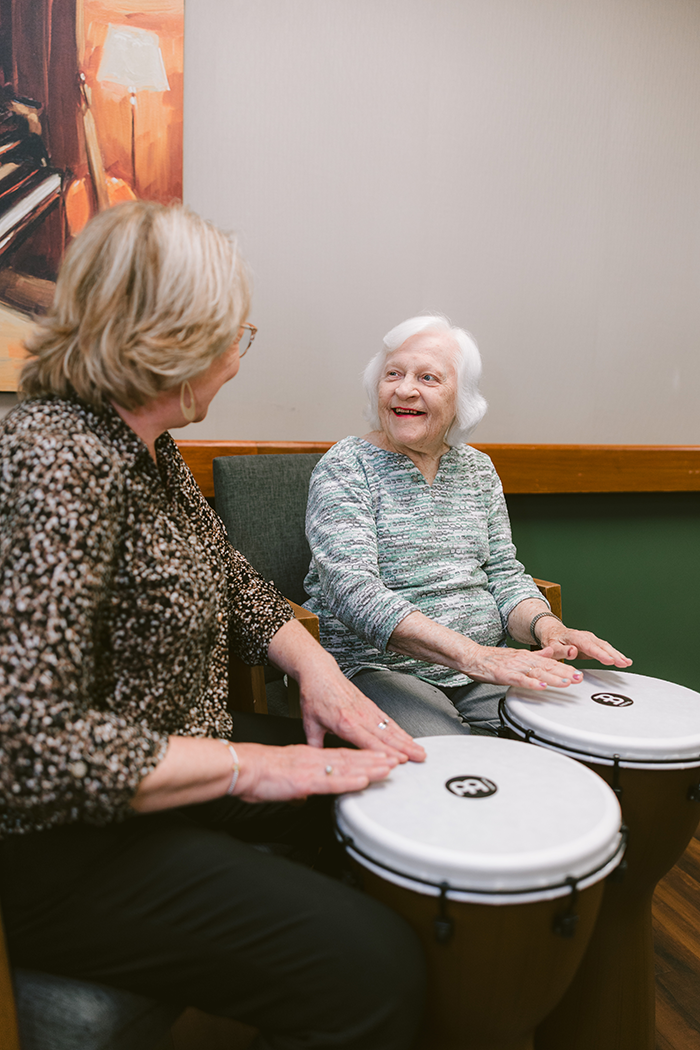 Older adult woman smiling while participating in a music therapy session at Agape Crossing. She plays hand drums alongside another woman in a comfortable indoor setting, fostering engagement, joy, and connection through rhythm and creative expression.