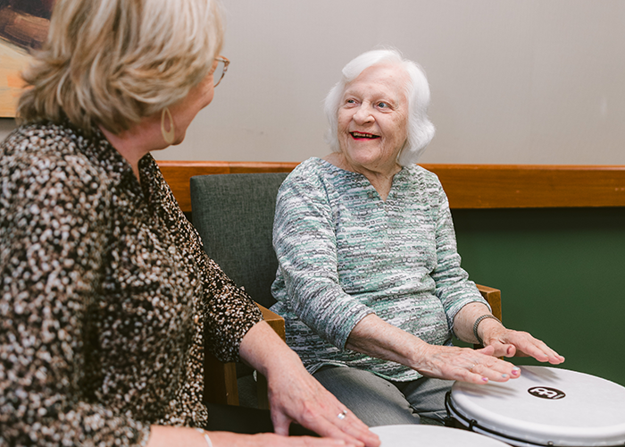 An older woman smiles while playing hand drums alongside another woman in a cozy indoor setting at Ohio Living Swan Creek's Agape Crossing. Both participants are seated and engaged in a music therapy activity that promotes joy, rhythm, and connection. A warm-toned painting and soft lighting create a welcoming environment designed to support emotional well-being and cognitive engagement for residents.