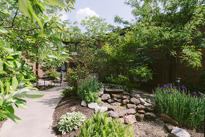 Serene garden courtyard at Agape Crossing, Ohio Living Breckenridge Village, featuring a stone water feature surrounded by lush greenery, flowering plants, and mature trees. A paved walkway winds through the landscaped area, offering benches and shaded spots for residents to relax and enjoy nature. The space reflects Ohio Living’s commitment to creating peaceful, therapeutic environments that support memory care and resident well-being.