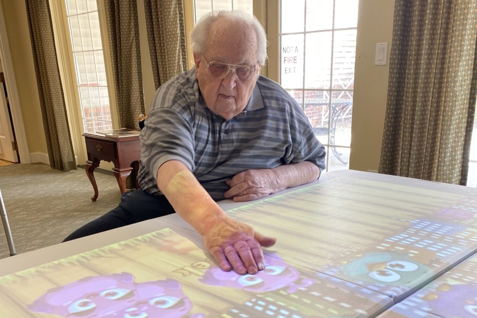 Older adult man engages with an interactive Lucynt projection table at Agape Crossing, part of Ohio Living’s memory care community. He touches colorful animated images displayed on the tabletop, participating in a sensory‑based activity designed to support cognitive stimulation, engagement, and joy for individuals living with dementia or Alzheimer’s.