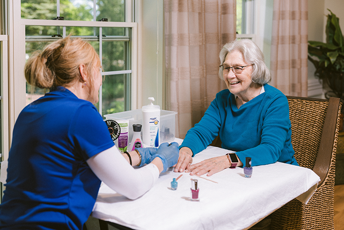 Older adult woman smiles while receiving a manicure from a caregiver at Agape Crossing, part of Ohio Living. The two sit at a table near large windows with natural light, surrounded by nail polish and lotion, creating a warm, relaxing environment that promotes personal care and connection for residents living with dementia or Alzheimer’s.
