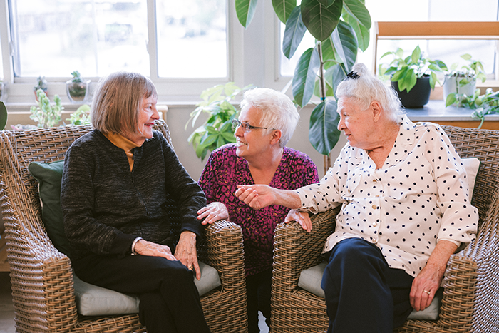 Three older adult women sit together in a bright, plant‑filled lounge at Agape Crossing, part of Ohio Living’s memory care community. They smile and chat warmly, creating a sense of friendship, comfort, and connection in a supportive environment designed for residents living with dementia or Alzheimer’s.