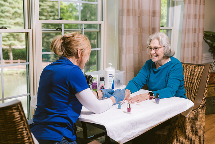 An older woman smiles while receiving a manicure from a caregiver in a bright, welcoming room at Agape Crossing, Ohio Living Rockynol. The caregiver, wearing a blue uniform, gently holds the resident’s hands across a table covered with a white cloth and nail care supplies. Sunlight streams through large windows overlooking a peaceful outdoor view, reflecting the community’s focus on personalized care, relaxation, and resident well-being.