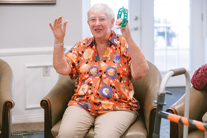 An older woman sits in a comfortable chair at Agape Crossing, Ohio Living Westminster-Thurber, smiling and raising her hands while holding a green tambourine. She wears a bright orange floral blouse and beige pants, participating in a lively music or movement activity in a well-lit community room. The setting reflects Ohio Living’s focus on joyful engagement, creativity, and supportive memory care experiences for residents.
