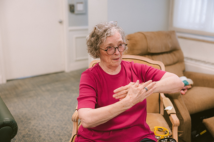    An older woman wearing glasses and a bright pink shirt sits comfortably in a cushioned chair at Agape Crossing, Ohio Living Westminster-Thurber. She smiles and gestures with her hands during a group activity or conversation in a cozy, well-lit room with neutral décor and soft furnishings. The setting reflects a warm, engaging environment designed to support memory care residents through social connection and meaningful interaction.