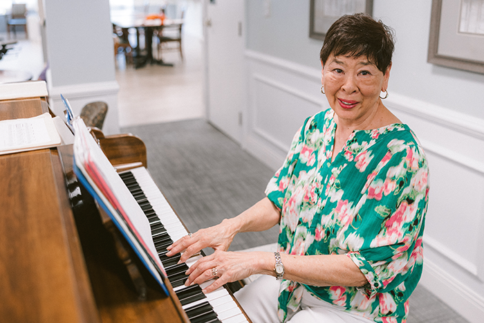 A volunteer at Agape Crossing, Ohio Living Westminster-Thurber, smiles while playing the piano in a bright, welcoming community space. She wears a green floral blouse and white pants, seated at a wooden piano with sheet music open in front of her. The setting reflects Ohio Living’s commitment to enriching memory care through music therapy, fostering joy, creativity, and emotional connection for residents.