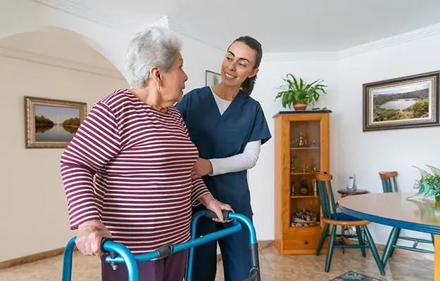 In‑home mobility assistance: Caregiver in blue scrubs helps an elderly woman using a walker in her living room, highlighting safety, independence, and supportive rehabilitation services.