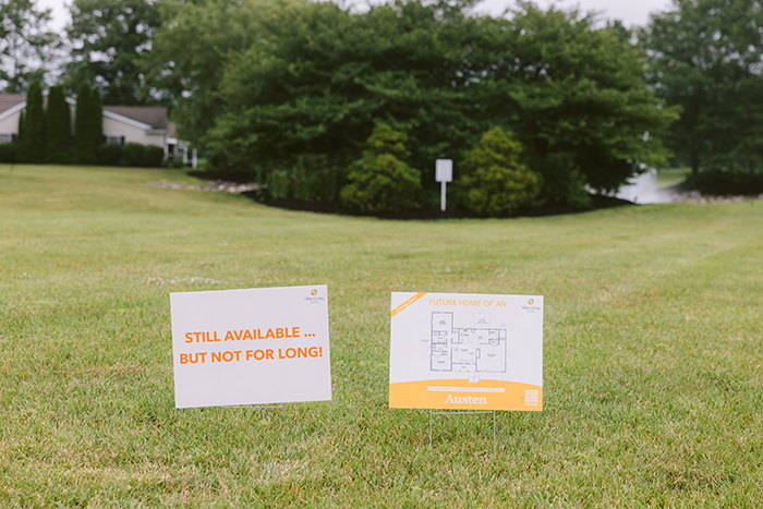 Two yard signs stand on a grassy lot at Ohio Living Cape May, promoting new senior living homes. The left sign reads “Still available … but not for long!” and the right sign displays a floor plan labeled “Future home of an Austen.” Behind the signs, trees, shrubs, and a pond create a peaceful residential backdrop, symbolizing upcoming housing opportunities and community growth for older adults.