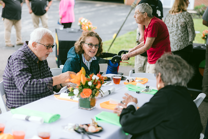 Future residents, residents and guests at Ohio Living Cape May enjoy a cheerful outdoor luncheon under a tent. Three older adults sit at a table decorated with bright orange and green napkins and a floral centerpiece of orange roses, smiling and conversing over refreshments. In the background, other attendees mingle near tables and a person with a walker joins the event, reflecting community connection and vibrant senior living engagement.