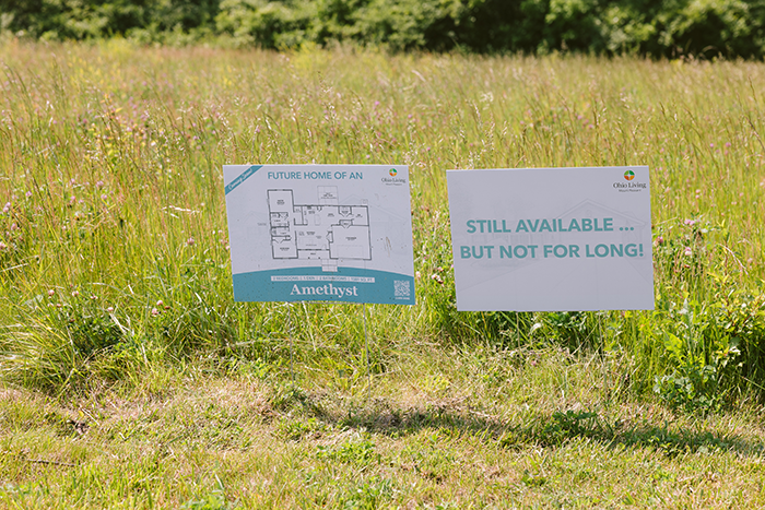 Two Ohio Living signs displayed in a grassy field with tall summer grass and wildflowers. The left sign shows a floor plan labeled “Future Home of an Amethyst,” while the right sign reads “Still available … but not for long!” Both feature the Ohio Living logo, promoting new senior living home development and availability.