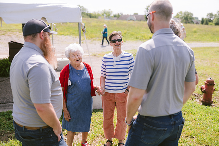 Group of people standing outdoors near a white tent, engaged in friendly conversation during an Ohio Living Mount Pleasant event. An older woman wearing a blue dress and red cardigan smiles while speaking with others dressed casually in gray shirts and jeans, and one woman in a striped top and orange pants. The sunny day and grassy field create a warm, welcoming community atmosphere.