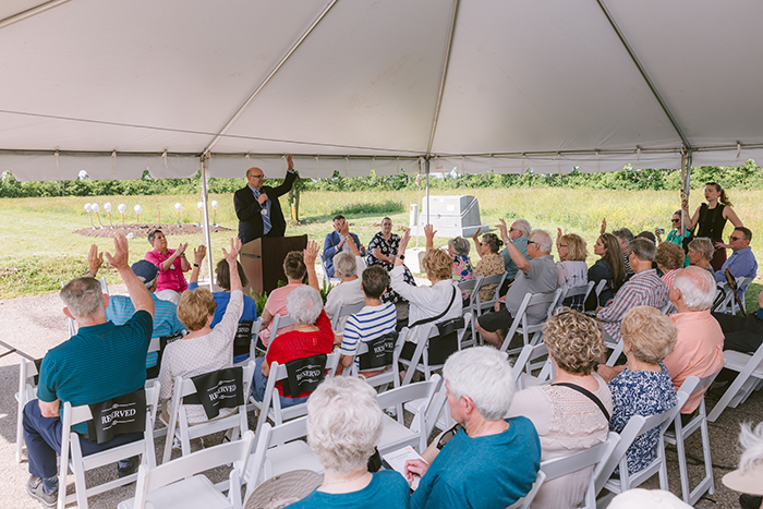Community members seated under a large outdoor tent listen to a speaker at a podium during an Ohio Living Mount Pleasant event. The audience, composed mostly of senior adults, raises their hands in participation while the speaker gestures in response. The setting features white folding chairs, a grassy field in the background, and a bright, sunny day—capturing engagement and connection within the senior living community.