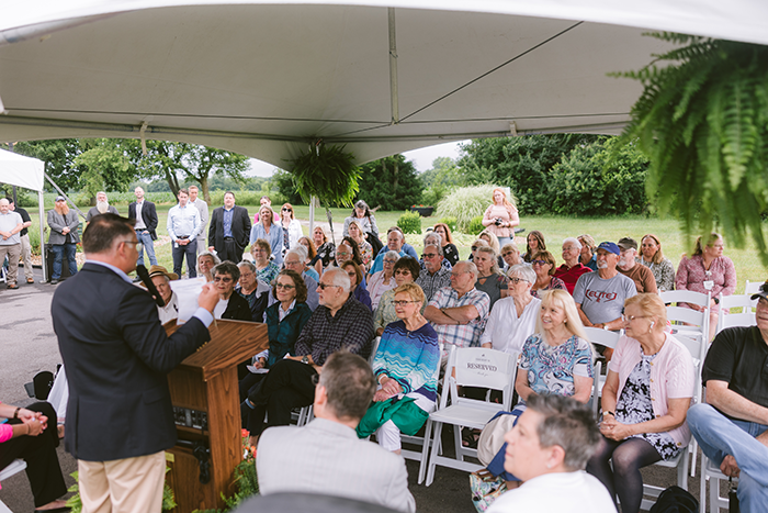 A speaker addresses a large group of attendees seated under a white canopy during an outdoor event at Ohio Living Cape May. The audience, composed mostly of older adults, listens attentively in rows of white chairs, with a “Reserved” sign visible near the front. Lush greenery and trees surround the gathering, creating a welcoming backdrop for the senior living community’s ceremony or celebration.