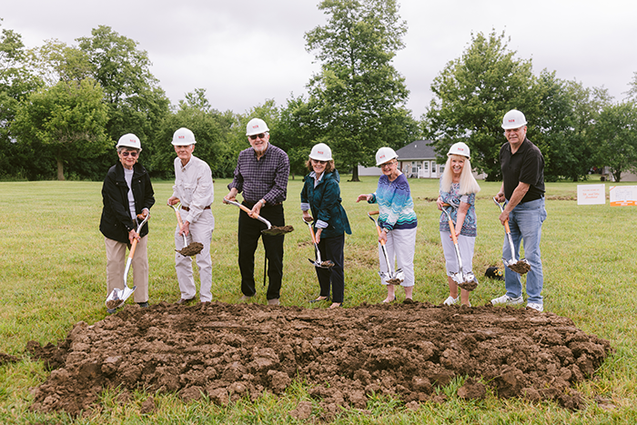 A group of seven people wearing white hard hats and holding shovels stand on a grassy field during a groundbreaking ceremony at Ohio Living Cape May. They smile as they lift soil, marking the start of a new development on campus. Trees and a residential building appear in the background under a cloudy sky, symbolizing community growth and progress in senior living.